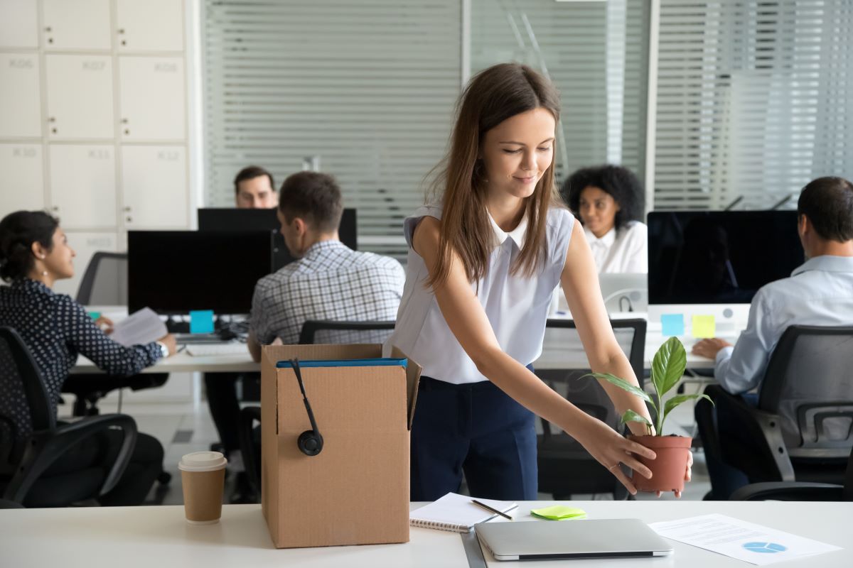 Smiling hired female company employee unpacking box with personal belongings at workplace on first working day in shared office